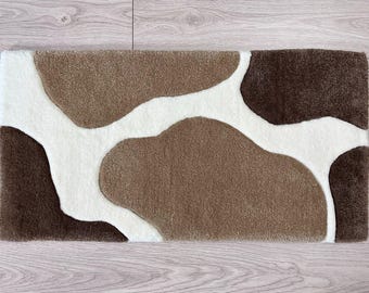 May include: A rectangular rug with a cowhide pattern. The rug features a design of brown, dark brown, and white irregular shapes. The rug is made of a soft, plush material, suitable for a living room or bedroom.