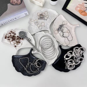 Op de afbeelding: Vijf ginkgo-blaadjesvormige keramische schalen in wit en zwart met gespikkelde afwerkingen. De schalen zijn gevuld met verschillende sieraden, waaronder kettingen, armbanden, ringen en een horloge.