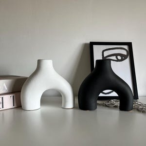 Può includere: Due vasi in ceramica a forma di arco. Un vaso è bianco e l'altro è nero. I vasi sono su una superficie bianca.