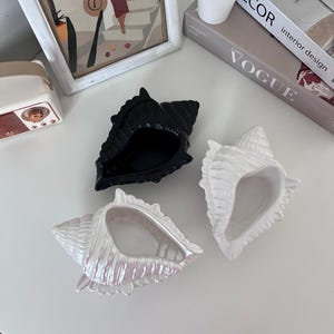 Puede incluir: Tres platos de cerámica con forma de concha. Uno es negro, otro es blanco y el último es blanco nacarado.