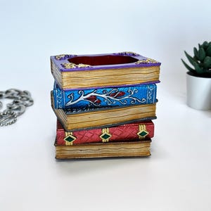 Puede incluir: Una pila de cuatro cajas decorativas con forma de libro. La caja superior es morada con detalles dorados, la segunda es azul con un diseño floral y la tercera es roja con formas de diamante verdes. Los libros están apilados uno encima del otro.
