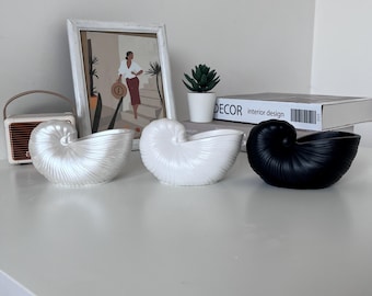 May include: Three ceramic seashell planters in white and black. The planters are on a white surface. The text "DECOR interior design" is visible on a book in the background.