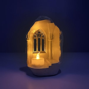 Puede incluir: Un portavelas blanco con forma de ventana gótica. El interior está iluminado con una cálida luz amarilla de una pequeña vela. La ventana tiene un diseño arqueado detallado, y el efecto general es sereno. Objeto decorativo.