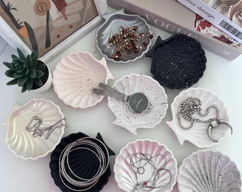 Puede incluir: Una colección de cuencos en forma de concha marina en varios colores, incluyendo rosa, blanco, negro y plateado. Algunos cuencos contienen joyas, un reloj y otros objetos pequeños. Una impresión artística enmarcada y libros decorativos están en el fondo.