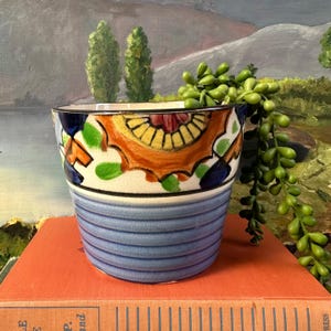 May include: A blue and white ceramic planter with a colorful floral design. The planter has a wide rim and a rounded base. It is decorated with a blue and white striped pattern around the bottom.