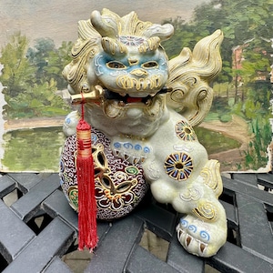 May include: A ceramic Foo Dog statue with a crackle glaze finish in cream, blue, and gold. The creature holds a black and gold sword in its mouth and rests a paw on a dark red, patterned sphere. A bright red tassel hangs from the sword handle.