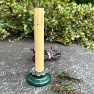 May include: A vintage electric candle lamp with a cream-colored candle and a green base. The candle has a textured appearance with simulated wax drips. The base is dark green with a silver-colored ruffled edge. A brown electrical cord is visible.
