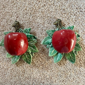 May include: Two ceramic apple wall decorations. Each apple is a glossy red color, set on a bed of green leaves. The leaves have a detailed, textured appearance. The stems are a dark brown color.