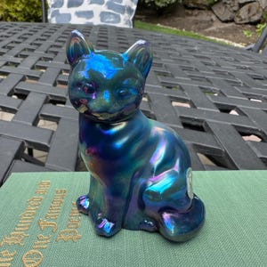 May include: A sitting cat figurine with a glossy, iridescent finish that shifts between deep blue, purple, and teal tones. The cat has pointed ears, alert eyes, and a smooth, reflective surface. A small circular sticker is visible on its side.