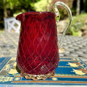 May include: A ruby red glass pitcher with a diamond pattern and a clear glass handle. The pitcher has a decorative gold-colored base and a spout for pouring. The pitcher is likely used for serving beverages.