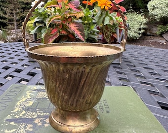 May include: A vintage brass planter with a twisted handle and a fluted body. The planter sits on a pedestal base and has a decorative rim. The planter is filled with colorful flowers. The planter is sitting on a green book.