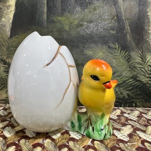 May include: A ceramic figurine featuring a white eggshell with gold-colored cracks and a yellow chick with an orange head. The chick is perched on green grass. The figurine is likely a decorative item.