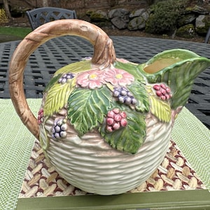 May include: A ceramic pitcher with a basket weave design, adorned with sculpted pink flowers, green leaves, and clusters of purple and red berries. The handle is a textured brown, and the spout is a light green.