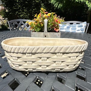 May include: An oval-shaped cream-colored ceramic planter featuring a textured exterior that mimics the appearance of a woven basket. The rim has a diagonal ribbed pattern, while the body displays a horizontal weave design with small raised details.
