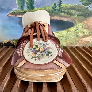 May include: A ceramic shoe planter with a brown and white glaze. The shoe has a floral design on the side and a brown suede lace.