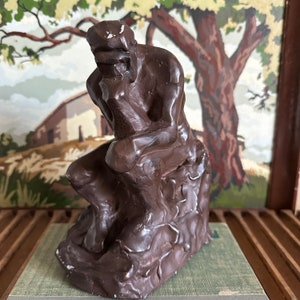 May include: A dark brown ceramic sculpture of a person sitting with their head in their hand, appearing to be in deep thought. The sculpture is set against a painted backdrop of a tree and a house.