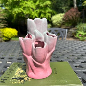 May include: A pink and green glazed ceramic tulip bud vase with four openings. The vase is sitting on a green book with the title "My Garden" and the author "Beebe Wilder".