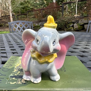 May include: A ceramic figurine of Dumbo the elephant from Disney's animated film Dumbo. The figurine is painted in shades of gray, pink, and yellow. Dumbo has large ears, a yellow hat, and a friendly expression.