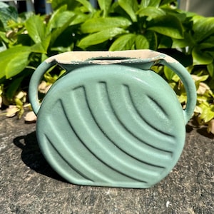 May include: A light green ceramic vase with two handles and a textured, curved design. The vase has a round shape and a distressed, off-white rim. The background features green foliage.