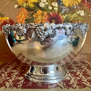 May include: A polished silver-toned metal bowl featuring a decorative rim adorned with raised, three-dimensional grape clusters and vine leaves. The bowl has a smooth, reflective surface and a sturdy, circular pedestal base.