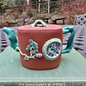 May include: A red clay teapot with a green bamboo handle and lid. The teapot is decorated with a green and white floral design and a round, green and white floral medallion. The teapot is sitting on a green book with the title "Trees & Landscapes".