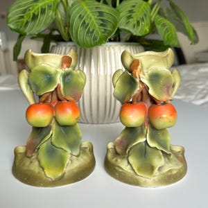 May include: A pair of ceramic candle holders shaped like cherry branches with green leaves and red cherries. The holders are in a vintage style and have a rustic look.