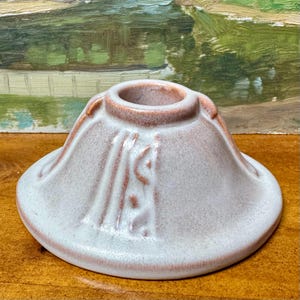May include: A ceramic candle holder with a wide base and a central opening. The holder is primarily a light pink color with accents of a darker pink. The design includes vertical lines and a textured surface.
