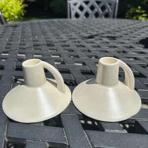 May include: Two off-white ceramic candle holders with a handle and a wide, shallow base. The candle holders are sitting on a dark gray table. The background is blurred, with greenery and a decorative metal chair visible.