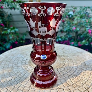May include: A ruby red glass vase with intricate etched designs. The vase features a bulbous base and a flared top, with detailed patterns of birds, buildings, and swirling motifs. The vase is approximately 8 inches tall.