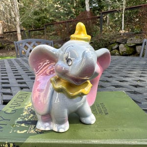 May include: A ceramic figurine of Dumbo the elephant from Disney's animated film Dumbo. The figurine is painted in shades of gray, pink, and yellow. Dumbo has large, pink ears and a yellow hat. The figurine is sitting on a book.