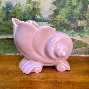May include: A light pink ceramic planter shaped like a conch shell. The vessel features a smooth, glossy finish with a spiraled design and a wide, open top. It stands on two curved, scroll-like feet that provide stability to the rounded base.