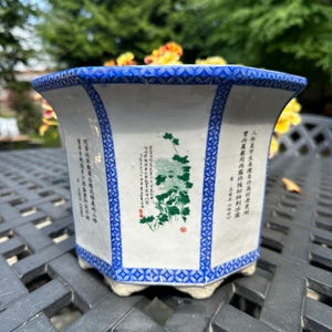 May include: A hexagonal ceramic planter with a white base and blue patterned borders. The sides feature green floral illustrations of chrysanthemums and columns of black Chinese calligraphy text with small red stamps.