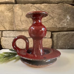 May include: A handcrafted, red ceramic candle holder with a circular base and a handle. The candle holder has a glossy finish and a unique design, perfect for adding a touch of elegance to any space. The handle is circular and adds to the overall aesthetic.