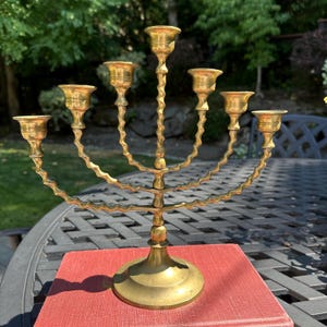 May include: A seven-branched brass candelabra featuring a central stem and six curved arms, each topped with a candle cup. The structure displays a twisted, spiral design on the vertical supports and a circular base for stability.