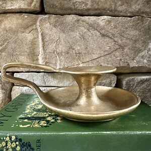 May include: A vintage brass candle holder with a handle. The candle holder has a round base and a central platform to hold a candle. The handle curves upwards. The candle holder is sitting on a green book.