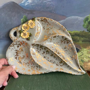 May include: A ceramic leaf-shaped serving dish with a gold speckled glaze and three raised sections. The dish is decorated with three yellow flowers with green leaves.