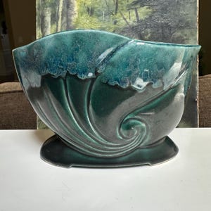May include: A ceramic planter with a deep teal and dark green glaze. The vessel features a wide, curved opening and a swirling, shell-like embossed pattern on the front. It sits on an oval base, showcasing a glossy finish with lighter highlights.