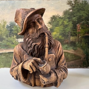 May include: A carved wooden bust of an elderly man with a long beard, hat, and pipe. The sculpture is a warm brown color with detailed carving, depicting the man's clothing and facial features. The background is a painted landscape.