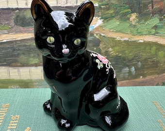 May include: A glossy black ceramic cat figurine in a seated position. The cat features bright green eyes, a small pink nose, and a delicate hand-painted floral design with pink flowers and green leaves on its back and side, finished with a smooth glaze.