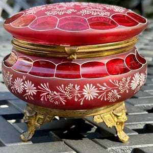 May include: A ruby red antique trinket box with a hinged lid and ornate gold-colored metal accents. The box features white floral designs and rests on decorative feet. The lid and sides have a geometric pattern.