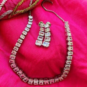 May include: A silver-toned necklace and matching earrings set. The jewelry features rectangular, clear stones set in a linear pattern. The set is displayed on a bright pink fabric, with a portion of an embroidered bag visible in the background.