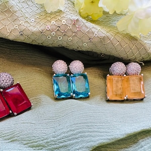 May include: Three pairs of gemstone earrings. One pair features red rectangular stones, another has blue rectangular stones, and the third pair has orange rectangular stones. Each earring has a round, sparkling top.