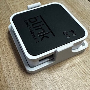 Può includere: Un modulo di sincronizzazione Blink Sync Module 2 bianco e nero con un'etichetta nera che dice "blink SYNC MODULE 2" e un logo nero che dice "blink".