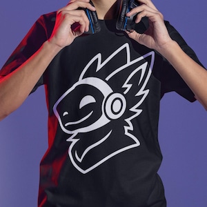 May include: Black t-shirt featuring a white graphic of a stylized creature's face. The design includes a circular element and jagged lines. The person in the image is holding headphones around their neck. The background is a gradient of purple and blue.