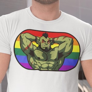 Puede incluir: Camiseta blanca con un personaje de orco de dibujos animados. El orco tiene piel verde, marcas rojas y barba negra. El orco está dentro de un óvalo de colores arcoíris. La camiseta se está usando.