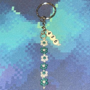 Puede incluir: Un llavero de cuentas verdes y blancas con un anillo plateado y un dije con los números 4, 5 y 6.