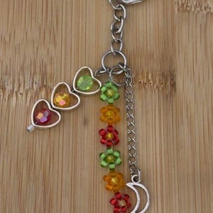 May include: A silver keyring with a decorative design. The keyring features three heart-shaped charms with colourful beads, a chain of flower-shaped beads, and a crescent moon charm. The beads are in shades of red, yellow, green, and orange.