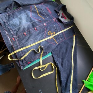 May include: A dark wash denim jacket with red contrast stitching, red buttons, and orange embroidered details. A yellow measuring tape is draped across the front and sleeve, showing measurements in inches up to approximately 29 inches.