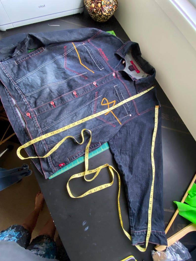 May include: A dark wash denim jacket with red contrast stitching, red buttons, and orange embroidered details. A yellow measuring tape is draped across the front and sleeve, showing measurements in inches up to approximately 29 inches.
