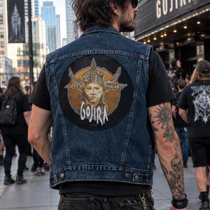 May include: A dark blue denim vest featuring a large circular patch on the back. The patch displays a graphic illustration of a figure wearing an ornate, horned headpiece against a circular gold background, with the text GOJIRA printed below.